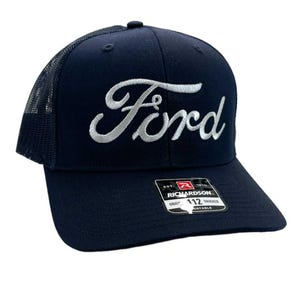 May include: Navy blue trucker hat with a mesh back and a curved brim. The word "Ford" is embroidered in white script across the front. A small tag with the Richardson logo is visible on the brim.