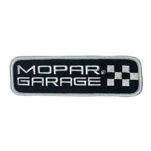 Puede incluir: Un parche rectangular negro con las palabras "MOPAR GARAGE" bordadas en blanco. Un diseño de bandera a cuadros está en el lado derecho. El parche tiene un borde plateado.