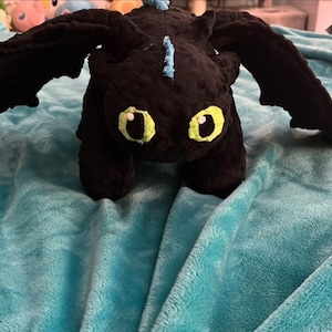May include: A black plush dragon toy with large black wings and bright yellow eyes. The dragon has light blue accents and is positioned on a turquoise blanket. The dragon is a soft toy.