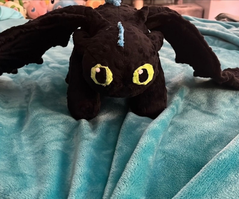 May include: A black plush dragon toy with large black wings and bright yellow eyes. The dragon has light blue accents and is positioned on a turquoise blanket. The dragon is a soft toy.