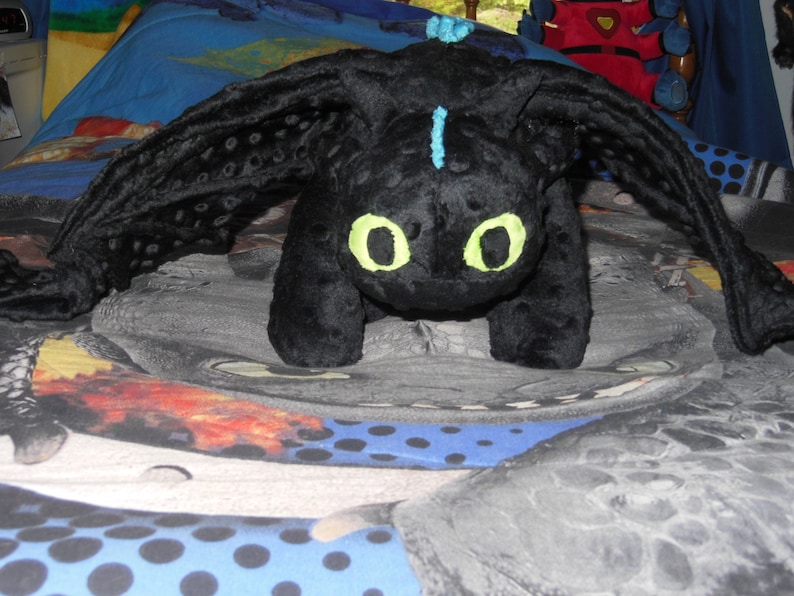May include: A black plush dragon toy with large, spread wings and bright yellow eyes. The dragon has a blue stripe down its head and is lying on a patterned bedspread. The toy is a soft, cuddly character.
