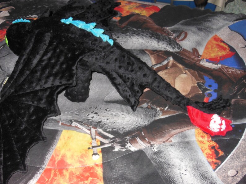 May include: A plush black dragon toy with blue spikes along its back and a red tail fin. The dragon has large wings and green eyes. The toy is lying on a patterned blanket.
