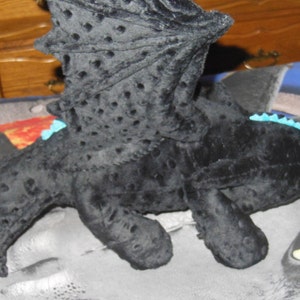 May include: A black plush dragon toy with textured wings and body. The dragon has green eyes, blue accents, and a detailed wing design. The toy is lying on a patterned surface.