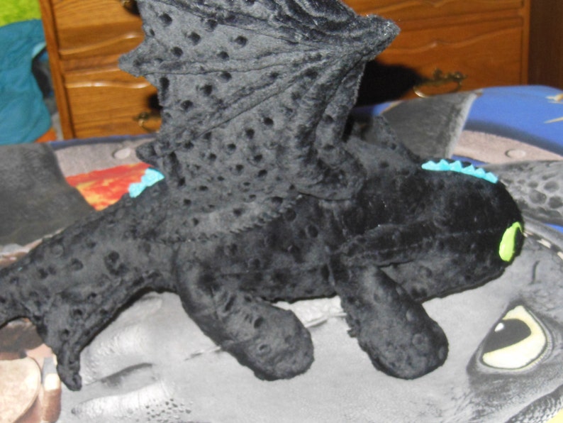 May include: A black plush dragon toy with textured wings and body. The dragon has green eyes, blue accents, and a detailed wing design. The toy is lying on a patterned surface.