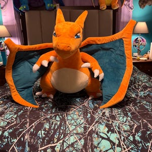 Pode incluir: Um grande peluche laranja da personagem Charizard com uma barriga cor de creme, membranas das asas azul-esverdeadas e garras brancas afiadas nas mãos e nos pés. A criatura está representada numa posição de pé com as asas bem abertas.
