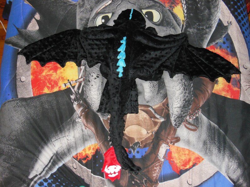 May include: A black dragon costume with large wings and a long tail. The costume has blue accents and is made of a soft, textured fabric. The costume is designed to resemble a dragon from the "How to Train Your Dragon" franchise.