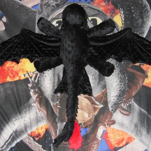 May include: A black plush dragon toy with large, textured wings and a red tail fin. The dragon is lying on a patterned background with images of dragons and flames.