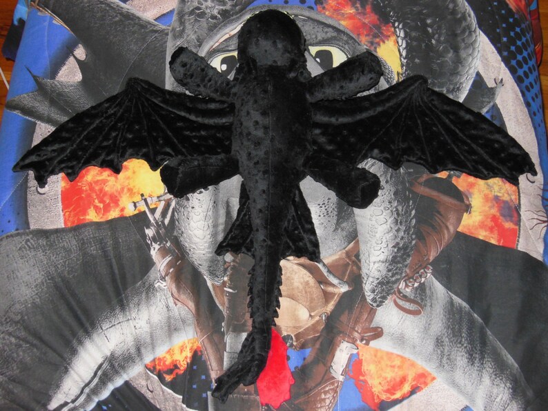 May include: A black plush dragon toy with large, textured wings and a red tail fin. The dragon is lying on a patterned background with images of dragons and flames.