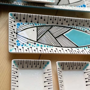 May include: A collection of white ceramic serving dishes featuring a fish design. The fish is outlined in black with geometric patterns and a turquoise accent. The set includes rectangular platters and square bowls, all with black and turquoise detailing.