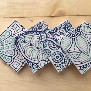 May include: Four square ceramic coasters with intricate hand-painted designs in teal and navy blue. Each coaster features a unique, symmetrical pattern on a white background. The coasters are arranged on a light wood surface.