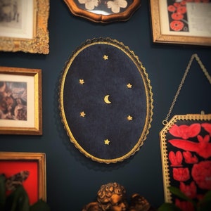 May include: An oval, dark blue velvet wall hanging with gold stars and a crescent moon. The hanging is framed with a gold lace-like trim. Other framed art pieces are visible in the background.