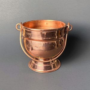 May include: A small, copper-colored cauldron with a rounded body and a handle. The cauldron has a small base and two small handles on either side. The handle is attached to the cauldron with a small loop. The cauldron is made of a shiny metal.