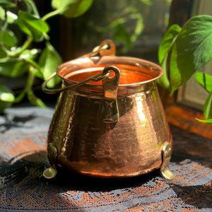 May include: A hammered copper cauldron with a polished finish, featuring a wide circular opening, a sturdy brass swing handle attached to two side mounts, and three curved brass feet that elevate the base from the surface below.
