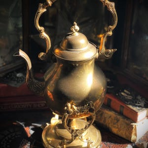 May include: A vintage brass teapot with ornate detailing, including a curved handle and decorative spout. The teapot sits on a matching stand with three feet. The image is lit with warm sunlight, highlighting the gold color and intricate design.
