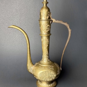 May include: A vintage brass pitcher with a long, curved spout and a handle. The pitcher is adorned with floral engravings and has a tall, ornate lid. The brass has a warm, golden hue, and the design suggests an antique or decorative item.