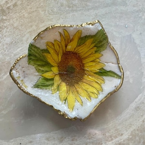 May include: A small, white, resin dish with a gold rim, featuring a painted sunflower with green leaves. The dish is signed "R. Rose" in the lower left corner.