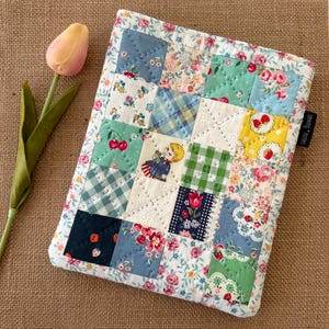 May include: A patchwork quilted case featuring squares of floral and patterned fabrics in blue, green, and pink. The case has a white floral border and the text "sewing notes".