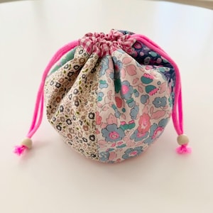 May include: A patchwork drawstring bag with a pink drawstring and wooden beads. The bag is made of floral patterned fabric in shades of pink, blue, and brown. The drawstring is bright pink, and the bag is sitting on a white surface.