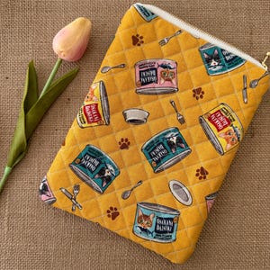 May include: Yellow quilted pouch with a zipper closure, featuring a repeating pattern of cat food cans, paw prints, and utensils. The cans have cat illustrations and Japanese text. A pink and yellow tulip is in the background.