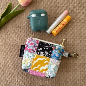 May include: A small, patchwork fabric pouch with a gold zipper and a small clasp. The pouch is filled with a lip balm, a tube of lip gloss, and a green case for wireless earbuds. A pink and yellow tulip is in the background.