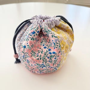 May include: A round drawstring bag with a patchwork design. The bag features floral patterns in pink, blue, and yellow. The drawstring is dark gray with a pink bead and tassel. The bag is approximately 6 inches in diameter.