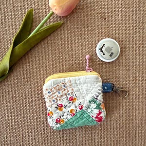 May include: A small, patchwork coin purse with a yellow zipper and a blue denim strap with a silver clasp. The purse features floral and geometric patterns in various colors. An Apple AirTag and a tulip are also in the image.