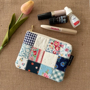May include: A patchwork cosmetic bag with a gold zipper, featuring floral and gingham patterns in various colors. The bag is next to a tulip and several beauty products, including mascara and eye drops.