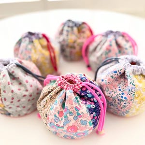 May include: A collection of small, round drawstring bags. Each bag is made from floral patterned fabric in various colors, including pink, blue, yellow, and gray. The bags have a drawstring closure with a contrasting pink or black cord.