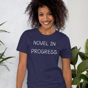 May include: A navy blue t-shirt with the white text "NOVEL IN PROGRESS". The shirt is made of a soft material and is being worn by a person. The person is smiling and is surrounded by green plants.