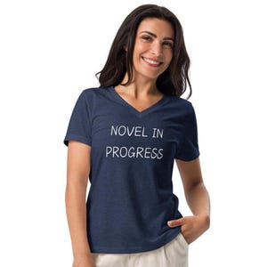 May include: A navy blue V-neck t-shirt with the white text "NOVEL IN PROGRESS". The shirt is made of a soft material and is suitable for casual wear. The model is smiling.
