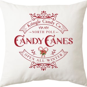 May include: White throw pillow with a red graphic design that says "Kringle Candy Co. North Pole Candy Canes Open All Winter". The graphic features a stylized candy cane and holly.