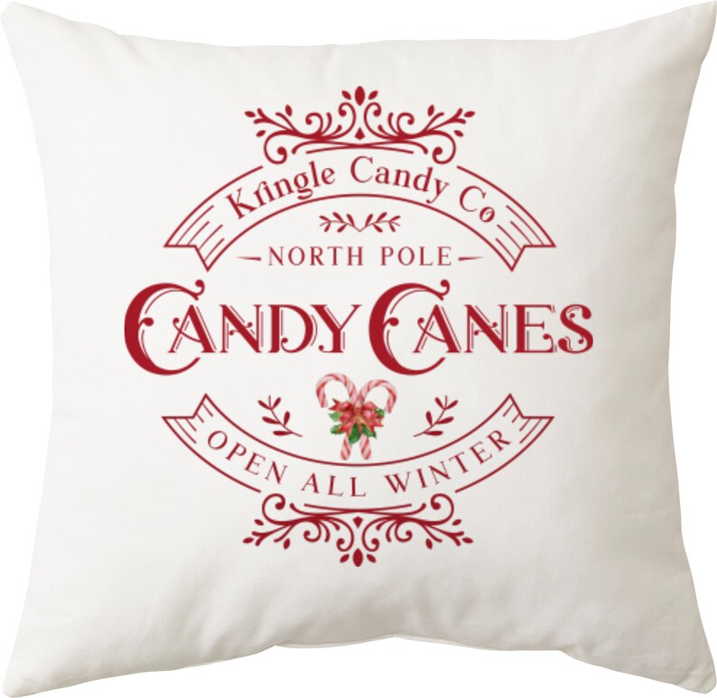 May include: White throw pillow with a red graphic design that says "Kringle Candy Co. North Pole Candy Canes Open All Winter". The graphic features a stylized candy cane and holly.