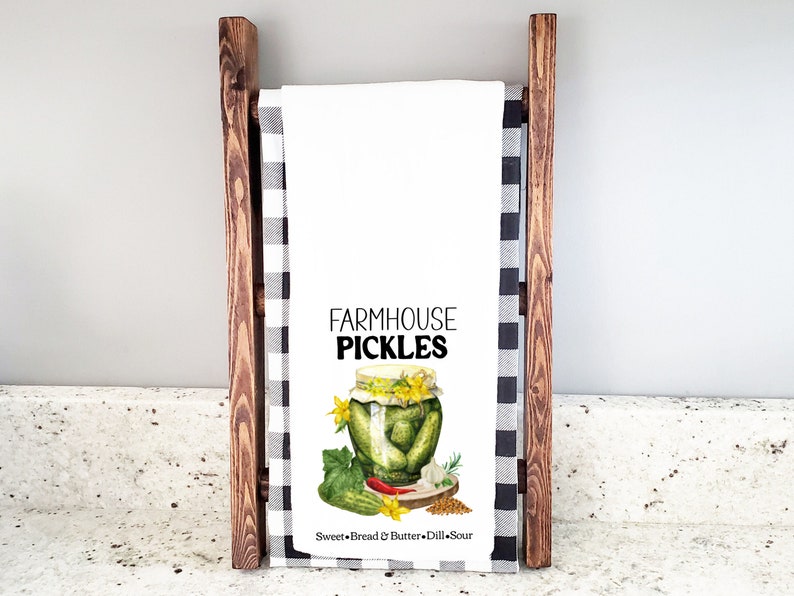 May include: White kitchen towel with a watercolor illustration of a jar of pickles. The text "FARMHOUSE PICKLES" is printed in black above the jar. The text "Sweet + Bread & Butter + Dill Sour" is printed in black below the jar.