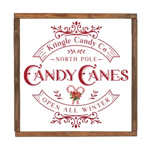 May include: A white sign with a red border and red text that reads "Kringle Candy Co. - North Pole - Candy Canes Open All Winter". The sign has a decorative border and a candy cane illustration.