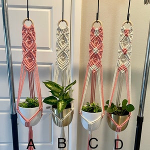 May include: Four macrame plant hangers with different color combinations of pink and white. Each hanger is holding a potted plant. The hangers are labeled A, B, C, and D.