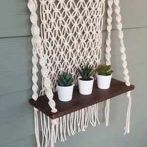 May include: A macrame wall hanging with a wooden dowel and a shelf underneath. The shelf is made of dark wood and has three small potted plants on it.
