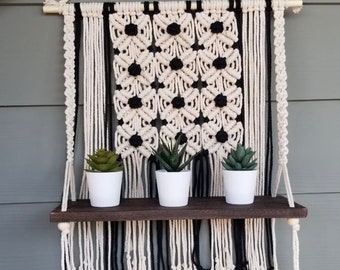 May include: A macrame wall hanging with a black and white geometric pattern and a wooden shelf with three small potted succulents.