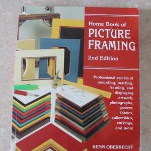Op de afbeelding: Een boek getiteld "Home Book of Picture Framing" met een rode kaft en de woorden "2nd Edition" gedrukt op de voorkant. De kaft van het boek toont een verscheidenheid aan fotolijsten in verschillende kleuren en stijlen, waaronder een hamer en een mes.