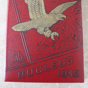 May include: A red leather-bound yearbook with a gold eagle design and the text "The Nucleus 1942" embossed on the front cover.
