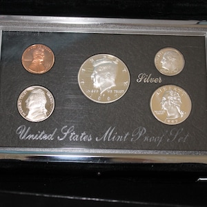 Könnte beinhalten: Ein 1997 United States Mint Silver Proof Set in einem rechteckigen Etui. Das Set enthält einen kupferfarbenen Cent, einen Nickel, einen Dime, einen Quarter und eine Half-Dollar-Münze. Der schwarze Einsatz trägt den Text United States Mint Proof Set.