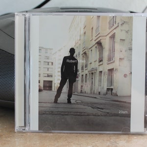 Può includere: Una custodia per CD con una foto in bianco e nero di una persona in piedi su una strada di città. Il testo "Robert Miles" e "23am" è stampato sulla custodia.