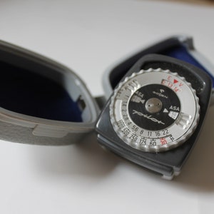 May include: A silver and black Gossen Pilot light meter with a black dial and red markings. The meter is in a gray case with a blue interior.