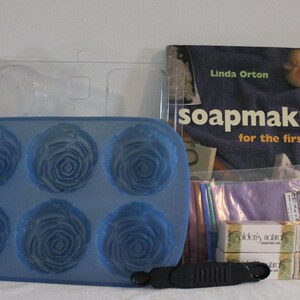 May include: A blue silicone mold with six rose-shaped cavities, a book titled Soapmaking for the First Time by Linda Orton, four small boxes of Golden's Naturals essential oils, a black plastic tool, and several colorful fabric strips.