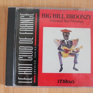以下が含まれることがあります： Le Hot Club De France Archive Seriesのアルバム「Big Bill Broonzy Unissued Test Pressings」のCDケース。カバーアートには、赤と白の背景にギターを弾くミュージシャンの水彩画イラストが描かれています。