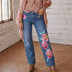 Könnte beinhalten: Hellblaue Jeans mit großen, auf der Vorderseite aufgemalten, rosa Blumendesigns. Die Jeans werden mit einem rosa Spitzentop, einem transparenten rosa Schal und mehrschichtigen Halsketten kombiniert. Das Model trägt braune Stiefeletten.