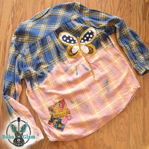 May include: A long-sleeved, plaid shirt in blue, yellow, and orange, with a butterfly applique. The shirt has a bleached effect on the lower half and a patch with floral and geometric patterns. The shirt is laid flat on a wooden surface.
