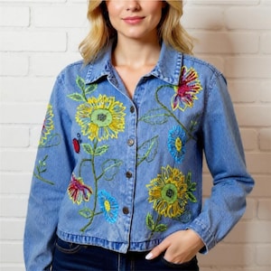 May include: Light blue denim jacket with embroidered sunflowers, blue and red flowers, and green leaves. The jacket has a button-up front and a collared neckline. The floral embroidery adds a colorful, artistic touch to the casual jacket.