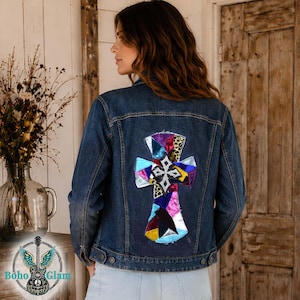 May include: A dark wash denim jacket with a colorful cross design on the back. The cross is made of various fabric patches in shades of blue, pink, purple, and leopard print. The jacket has a classic button-up design.