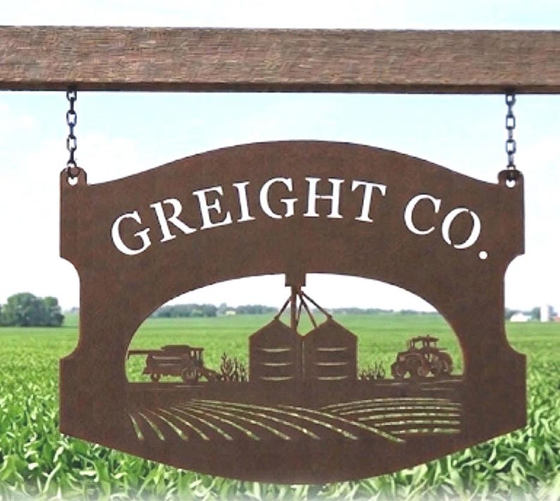 May include: A brown metal sign hanging from chains with the text GREIGHT CO. in white lettering. The sign features a cutout design depicting a farm scene with a combine harvester, grain silos, a tractor, and rows of crops in a field.