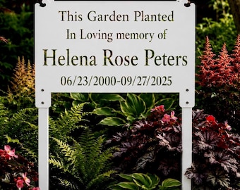 May include: A rectangular white metal garden sign with decorative scroll corners, mounted on two vertical white stakes. The sign features black text reading: This Garden Planted In Loving memory of Helena Rose Peters 06/23/2000-09/27/2025.